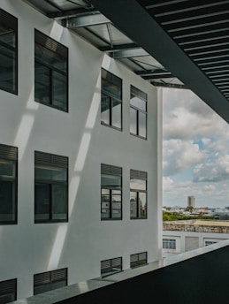 A modern building facade with several large windows under a slatted metal roof. Sunlight casts shadows across the white exterior wall. In the background, there is a view of a cityscape with a tall building, trees, and a cloudy sky.