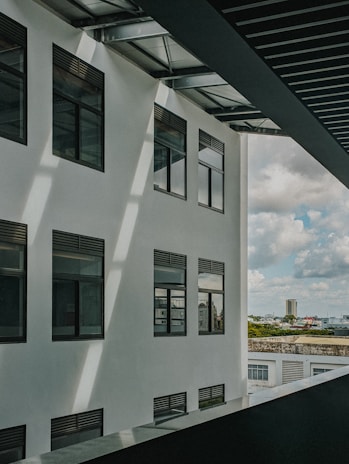 A modern building facade with several large windows under a slatted metal roof. Sunlight casts shadows across the white exterior wall. In the background, there is a view of a cityscape with a tall building, trees, and a cloudy sky.