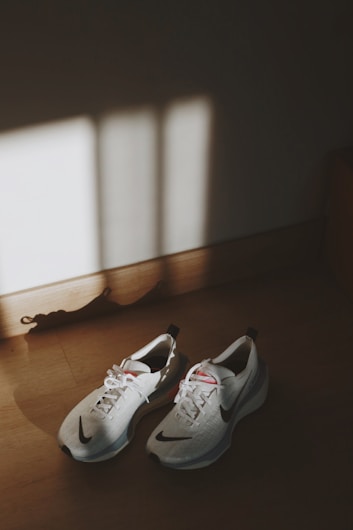 A pair of white athletic shoes with black accents is placed on a wooden floor. Sunlight streams through a window, casting shadows on the floor and across the sneakers.