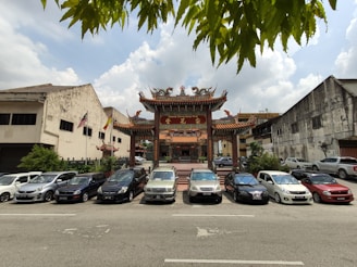 A traditional Chinese temple with ornate architecture and intricate designs, surrounded by parked cars in an urban setting. Flags are visible, and the surrounding buildings appear worn and rustic. Leaves partially frame the top of the image.
