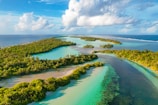 Aerial view of a tropical island chain surrounded by vibrant turquoise waters. Lush greenery and palm trees cover the islands. The ocean extends into the horizon under a sky filled with clouds.