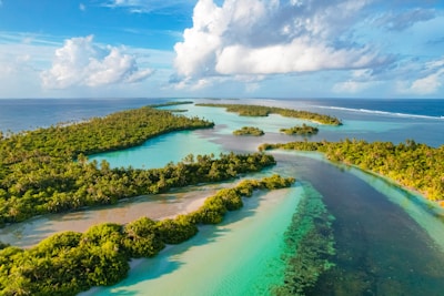 Aerial view of a tropical island chain surrounded by vibrant turquoise waters. Lush greenery and palm trees cover the islands. The ocean extends into the horizon under a sky filled with clouds.