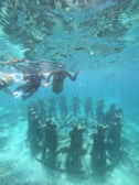 A group of people snorkeling above an underwater sculpture of human figures arranged in a circle. The sculptures are submerged in clear blue water, allowing a view of the sea floor and marine life. The scene depicts a blend of art and nature.