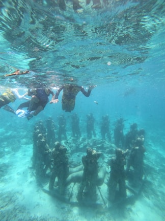 A group of people snorkeling above an underwater sculpture of human figures arranged in a circle. The sculptures are submerged in clear blue water, allowing a view of the sea floor and marine life. The scene depicts a blend of art and nature.