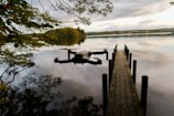 A drone hovers above a calm lake near a wooden pier that extends into the water. The sky is overcast, casting soft reflections on the lake's surface. Trees with autumn foliage partially frame the scene, adding a touch of color.
