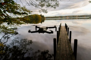 A drone hovers above a calm lake near a wooden pier that extends into the water. The sky is overcast, casting soft reflections on the lake's surface. Trees with autumn foliage partially frame the scene, adding a touch of color.
