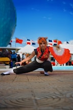 A person is dressed in a costume with an orange wig, crouching in a dynamic pose. They are holding a red and white fan in one hand. In the background, there are flags from multiple countries and tents, suggesting an outdoor event or festival.