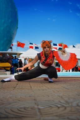 A person is dressed in a costume with an orange wig, crouching in a dynamic pose. They are holding a red and white fan in one hand. In the background, there are flags from multiple countries and tents, suggesting an outdoor event or festival.