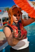 A person dressed in a vibrant cosplay outfit featuring a red and black sleeveless top with golden patterns, wearing elaborate face paint and ears resembling an animal. The individual has bright orange hair and is holding a colorful fan with orange and white patterns. The background includes a pool area and blurred tents.