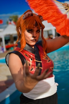 A person dressed in a vibrant cosplay outfit featuring a red and black sleeveless top with golden patterns, wearing elaborate face paint and ears resembling an animal. The individual has bright orange hair and is holding a colorful fan with orange and white patterns. The background includes a pool area and blurred tents.