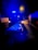An ambulance is parked on a road at night with its blue emergency lights flashing. In the foreground, a stretcher covered with a blue sheet is visible under the intense lighting. The surrounding area appears dimly lit, contrasting with the bright lights of the ambulance.