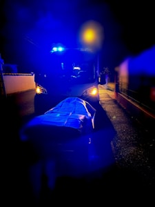 An ambulance is parked on a road at night with its blue emergency lights flashing. In the foreground, a stretcher covered with a blue sheet is visible under the intense lighting. The surrounding area appears dimly lit, contrasting with the bright lights of the ambulance.