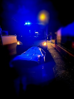 An ambulance is parked on a road at night with its blue emergency lights flashing. In the foreground, a stretcher covered with a blue sheet is visible under the intense lighting. The surrounding area appears dimly lit, contrasting with the bright lights of the ambulance.