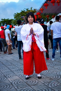 A person dressed in a traditional Japanese outfit with a white top and red hakama, holding prayer beads, stands in the center of a tiled outdoor area. Around them, there are people casually dressed, some with their backs turned. Red lanterns hang above, suggesting a cultural festival atmosphere. Trees and a cloudy sky are visible in the background.