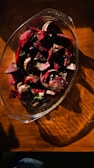 A glass dish filled with chopped red beets, garnished with small green herbs, sits on a wooden table. The lighting creates dramatic shadows and highlights across the surface.