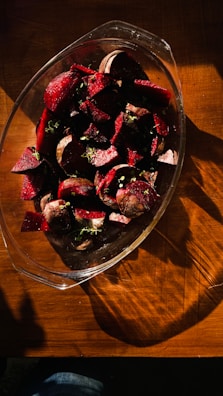 A glass dish filled with chopped red beets, garnished with small green herbs, sits on a wooden table. The lighting creates dramatic shadows and highlights across the surface.