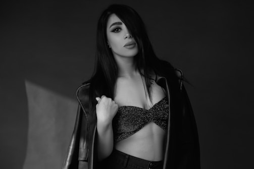 A woman with long dark hair is posing against a plain dark background. She is wearing a fitted, sparkly top and a leather jacket draped over her shoulders. Her expression is serious and confident, with direct eye contact.