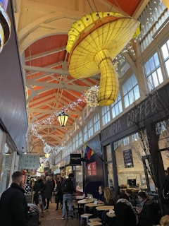 A bustling indoor marketplace with high ceilings and exposed wooden beams, featuring a large, decorative mushroom-shaped light fixture. There are various shops and cafes lining the walkway, with people sitting at tables and walking through the corridor. Festive string lights hang above, adding a lively and cozy atmosphere.