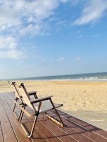Two folding chairs with wooden frames and light fabric seats are placed on a wooden deck overlooking a sandy beach with gentle waves in the background. The sky is mostly clear with a few scattered clouds.