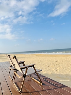 Two folding chairs with wooden frames and light fabric seats are placed on a wooden deck overlooking a sandy beach with gentle waves in the background. The sky is mostly clear with a few scattered clouds.
