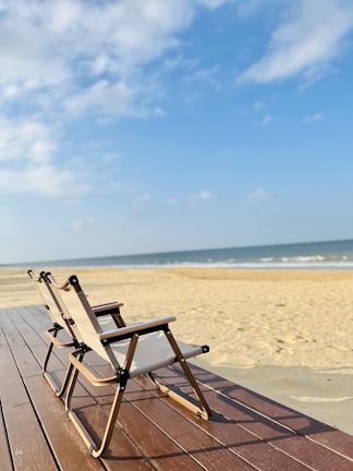 Two folding chairs with wooden frames and light fabric seats are placed on a wooden deck overlooking a sandy beach with gentle waves in the background. The sky is mostly clear with a few scattered clouds.