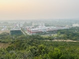 A sprawling campus with multiple buildings surrounded by lush greenery. The architecture features a mix of modern and traditional designs, with a prominent central building flanked by others. The horizon shows a hazy skyline with distant hills under a serene sky.
