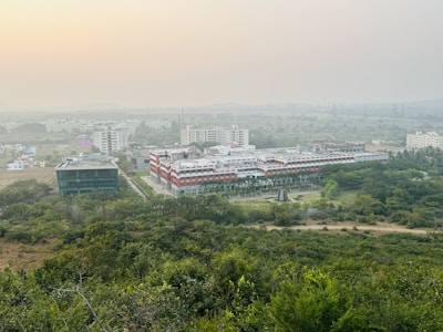 A sprawling campus with multiple buildings surrounded by lush greenery. The architecture features a mix of modern and traditional designs, with a prominent central building flanked by others. The horizon shows a hazy skyline with distant hills under a serene sky.