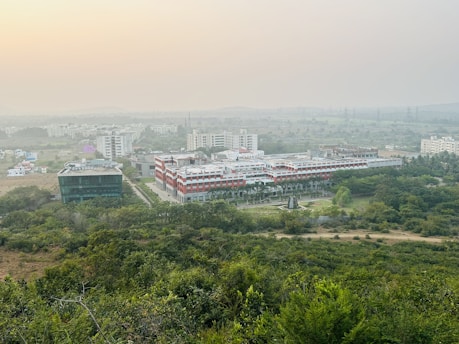 A sprawling campus with multiple buildings surrounded by lush greenery. The architecture features a mix of modern and traditional designs, with a prominent central building flanked by others. The horizon shows a hazy skyline with distant hills under a serene sky.