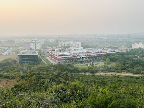 A sprawling campus with multiple buildings surrounded by lush greenery. The architecture features a mix of modern and traditional designs, with a prominent central building flanked by others. The horizon shows a hazy skyline with distant hills under a serene sky.