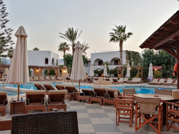 A luxurious outdoor pool area surrounded by numerous lounge chairs and umbrellas. The setting includes palm trees, a well-maintained garden, and a building with an arched facade in the background. The environment suggests a serene and relaxing atmosphere, indicative of a resort or high-end residential space.