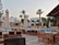 A luxurious outdoor pool area surrounded by numerous lounge chairs and umbrellas. The setting includes palm trees, a well-maintained garden, and a building with an arched facade in the background. The environment suggests a serene and relaxing atmosphere, indicative of a resort or high-end residential space.