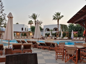A luxurious outdoor pool area surrounded by numerous lounge chairs and umbrellas. The setting includes palm trees, a well-maintained garden, and a building with an arched facade in the background. The environment suggests a serene and relaxing atmosphere, indicative of a resort or high-end residential space.