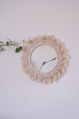 A round mirror is adorned with a beige macrame fringe border, hanging on a plain light-colored wall. A delicate green vine with a few leaves is draped over one side of the mirror, adding a touch of nature to the minimalist decor.
