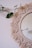 A round macramé mirror with intricate braided and fringed patterns is hanging on a plain wall. A green vine with leaves drapes across the top left corner, adding a touch of nature to the scene.