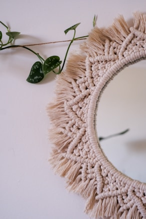 A round macramé mirror with intricate braided and fringed patterns is hanging on a plain wall. A green vine with leaves drapes across the top left corner, adding a touch of nature to the scene.