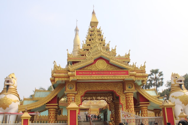 An ornate, gold-adorned temple entrance with intricate details, featuring multiple tiers and spires. Flanking the entrance are two large lion statues. In the background, a tall golden pagoda is visible. The scene is surrounded by palm trees and a clear sky.