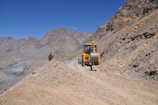A construction vehicle is positioned on a rocky dirt road in a mountainous area. The vehicle is yellow and appears to be moving or parked on the pathway. A man wearing dark clothing is walking nearby. The surrounding landscape is arid, with towering, rugged mountains and a clear blue sky.