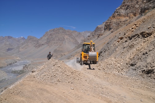 A construction vehicle is positioned on a rocky dirt road in a mountainous area. The vehicle is yellow and appears to be moving or parked on the pathway. A man wearing dark clothing is walking nearby. The surrounding landscape is arid, with towering, rugged mountains and a clear blue sky.
