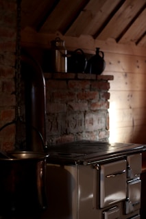 A rustic kitchen setting with a vintage wood-burning stove positioned against a brick wall, featuring a metal chimney pipe extending upwards. On a wooden shelf above the stove, there are several metal pots and a tin canister. Soft, ambient light illuminates part of the wooden wall and highlights the texture of the bricks.