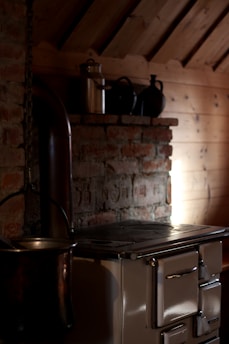 A rustic kitchen setting with a vintage wood-burning stove positioned against a brick wall, featuring a metal chimney pipe extending upwards. On a wooden shelf above the stove, there are several metal pots and a tin canister. Soft, ambient light illuminates part of the wooden wall and highlights the texture of the bricks.