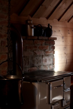 A rustic kitchen setting with a vintage wood-burning stove positioned against a brick wall, featuring a metal chimney pipe extending upwards. On a wooden shelf above the stove, there are several metal pots and a tin canister. Soft, ambient light illuminates part of the wooden wall and highlights the texture of the bricks.