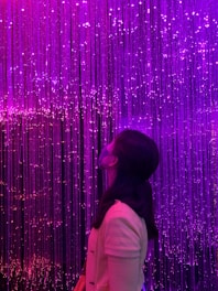 A woman stands looking upwards in front of an illuminated backdrop of cascading fiber optic lights. The lights create a vibrant, glowing effect in shades of pink and purple, giving a sense of a surreal or dreamlike atmosphere.