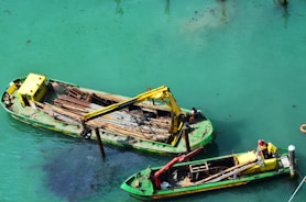 Two green and yellow barges are situated on turquoise water. Each barge is equipped with cranes, and workers can be seen on board. The barges carry long metal rods, and tools are visible, indicating construction or maintenance work in progress. The clear water highlights the dusty clouds formed by underwater activity.