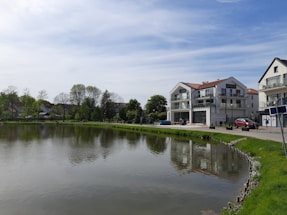 A tranquil lakeside scene with modern residential buildings and a clear sky. The water reflects the architecture and surrounding greenery. Trees line the distant background, adding a natural touch to the urban setting.