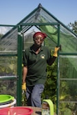 A person wearing a red headscarf and yellow gloves stands in the doorway of a small green greenhouse. They are dressed in a dark green polo shirt and gray pants. The greenhouse is surrounded by lush green plants and the sky is clear and blue. In the foreground, there are baskets and a bottle labeled Liquid Seaweed on a table.