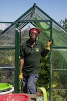 A person wearing a red headscarf and yellow gloves stands in the doorway of a small green greenhouse. They are dressed in a dark green polo shirt and gray pants. The greenhouse is surrounded by lush green plants and the sky is clear and blue. In the foreground, there are baskets and a bottle labeled Liquid Seaweed on a table.