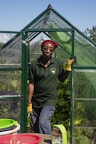 A person wearing a red headscarf and yellow gloves stands in the doorway of a small green greenhouse. They are dressed in a dark green polo shirt and gray pants. The greenhouse is surrounded by lush green plants and the sky is clear and blue. In the foreground, there are baskets and a bottle labeled Liquid Seaweed on a table.