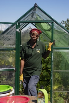 A person wearing a red headscarf and yellow gloves stands in the doorway of a small green greenhouse. They are dressed in a dark green polo shirt and gray pants. The greenhouse is surrounded by lush green plants and the sky is clear and blue. In the foreground, there are baskets and a bottle labeled Liquid Seaweed on a table.