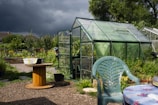 A green greenhouse stands prominently in a lush garden, surrounded by various plants and shrubs. In the foreground, there is a plastic chair and a wooden spool used as a table. A dark, stormy sky looms in the background, creating a stark contrast with the sunlit greenery.