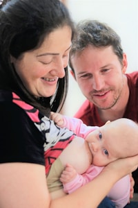 A woman is breastfeeding a baby while smiling. A man is leaning in close, looking at the baby with a happy expression. The baby is wearing a pink onesie.